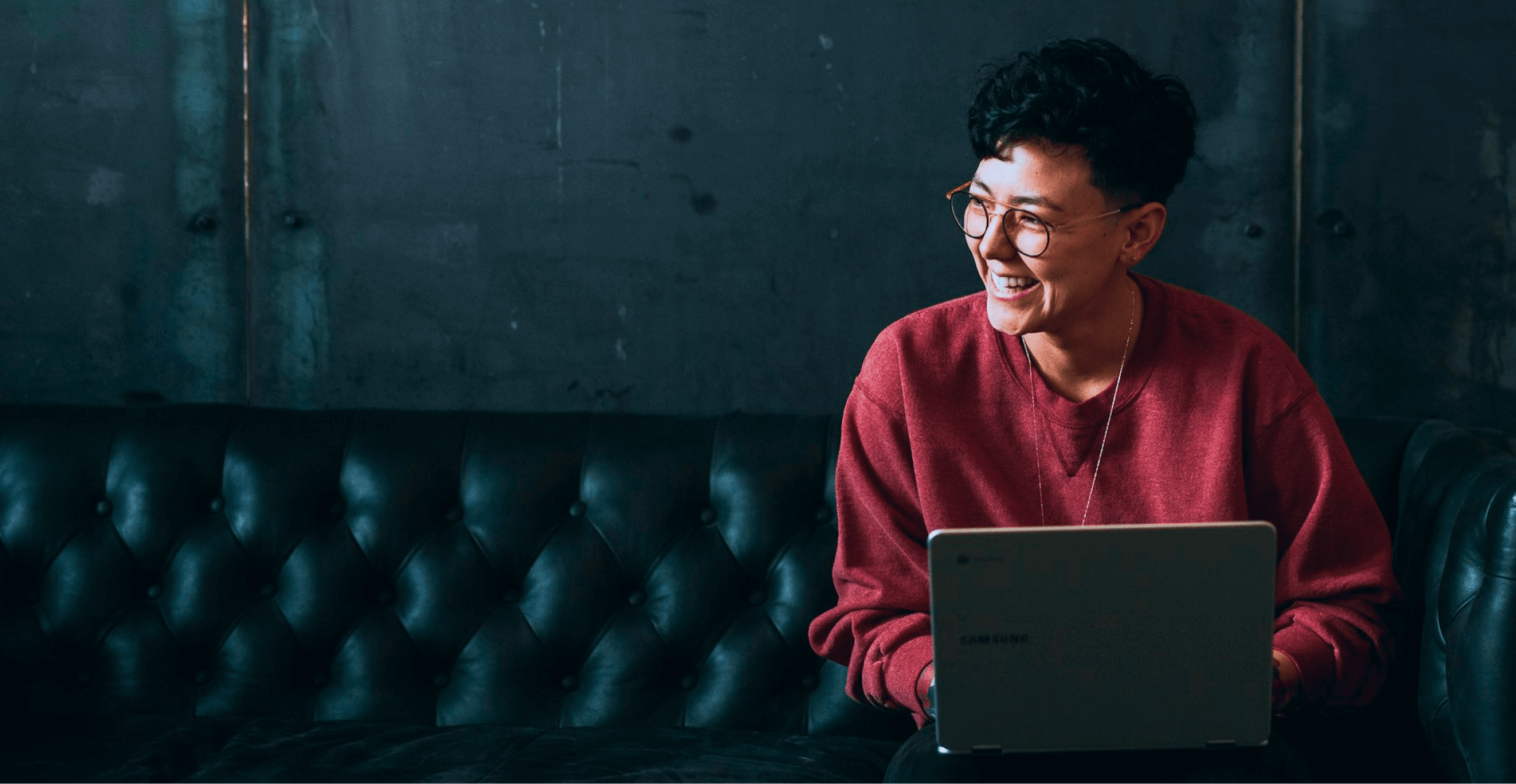 A person wearing glasses and a red sweater sitting on a couch with their laptop in their lap.